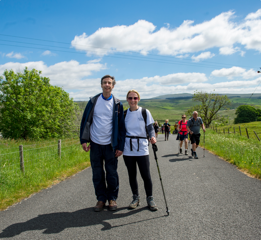 A couple pose for a photo in lush green countryside, it's a bright day. They are smiling, wearing a Bubble branded shirt. Other hikers march forward, on the same road behind them.